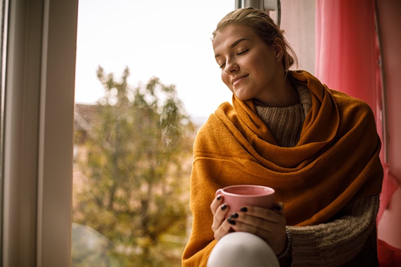 5 Tips to Improve Your Indoor Air Quality This Fall. Image of a woman sitting by a window drinking a hot beverage and enjoying the outdoor fall view.
